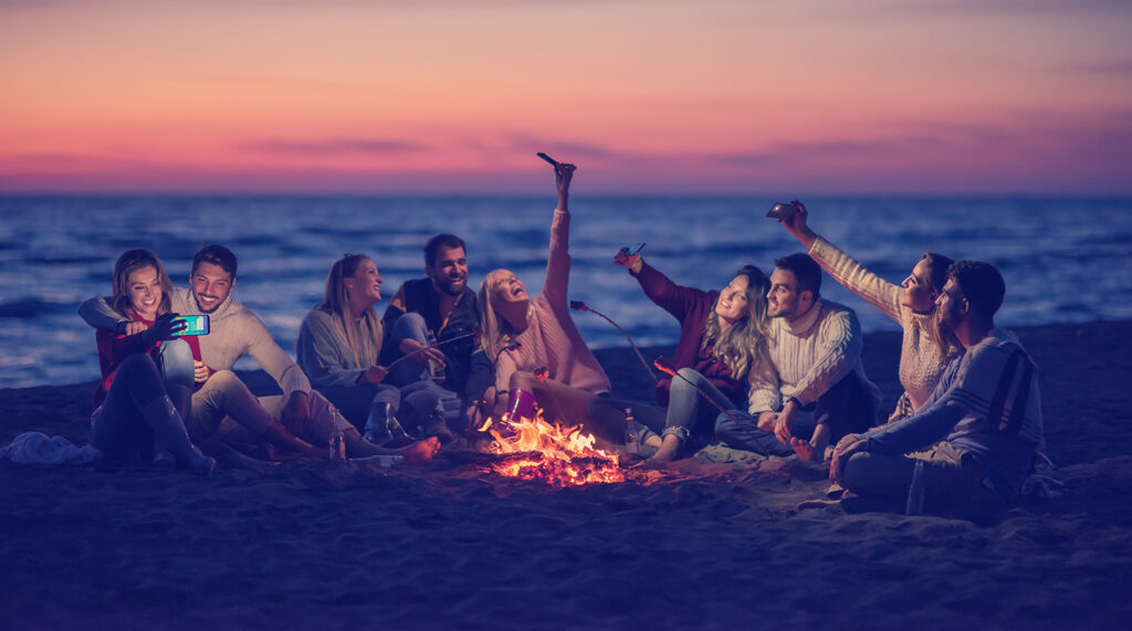 A diverse group of friends enjoy a bonfire on the beach at sunset.