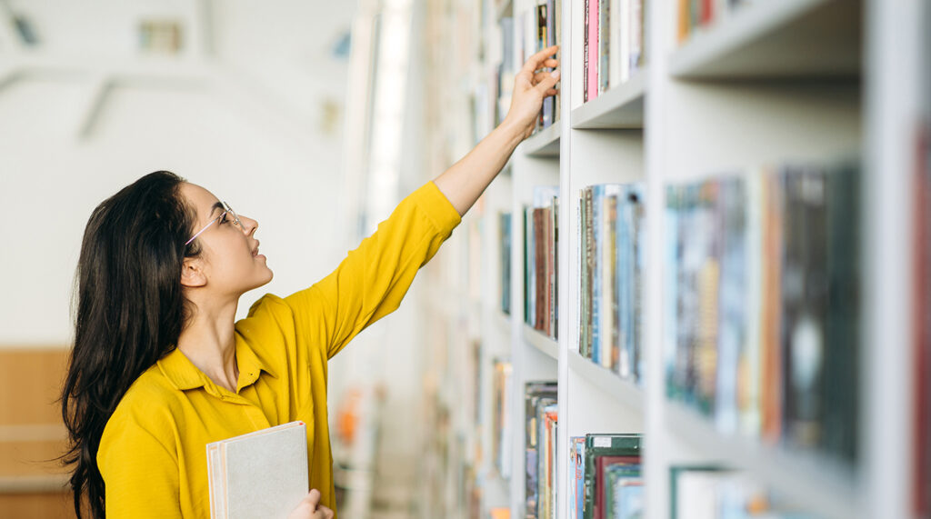 Young woman reaches for a book on a library shelf